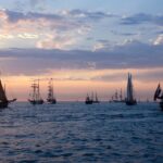 A fleet of classic sailing ships on the open sea during a stunning sunset, offering a serene maritime scene.